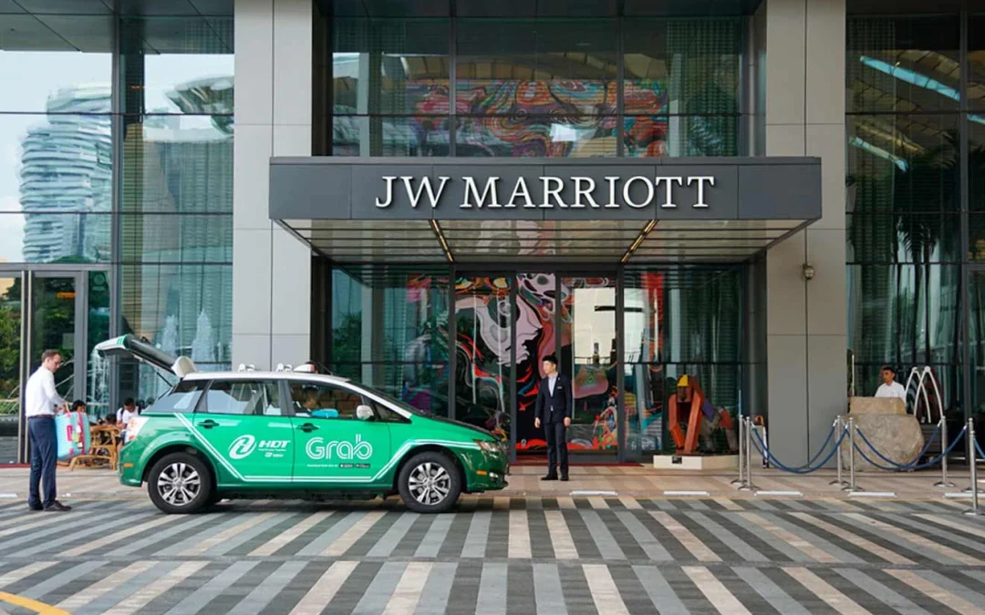 A green Grab taxi is parked outside the entrance of the JW Marriott hotel, with hotel staff assisting a guest at the entrance.