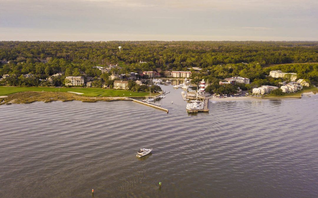 Aerial view of a waterfront town with a marina, surrounded by dense green trees. A yacht is sailing in the water towards the marina, structures and boats are visible on the shoreline.